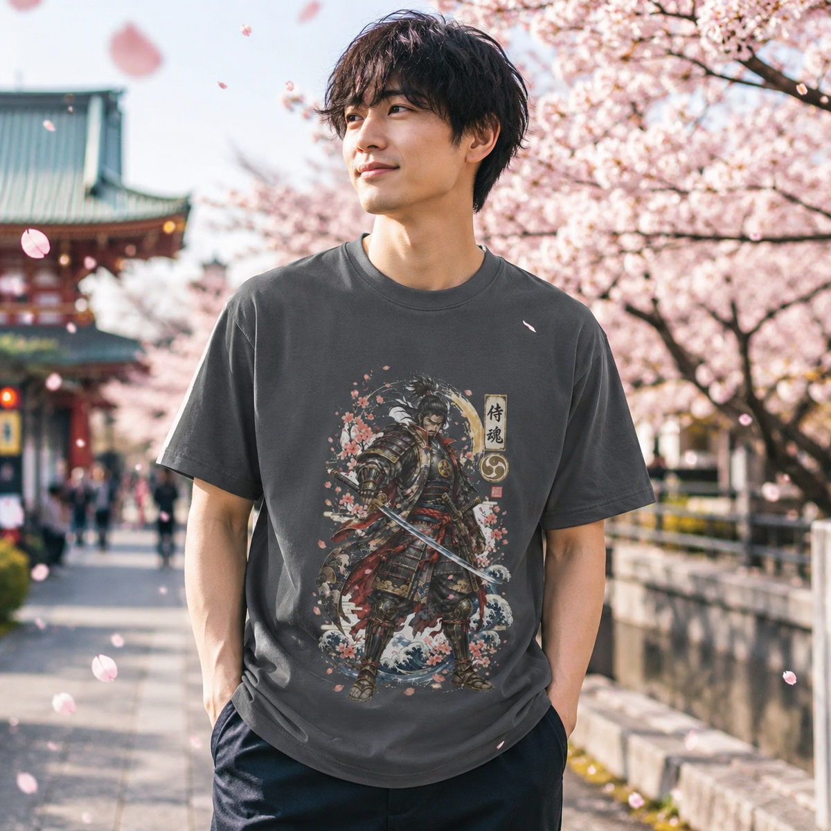 A man wearing a dark gray graphic t-shirt with a samurai warrior print, standing outdoors in front of blooming cherry blossom trees and a traditional Japanese temple.