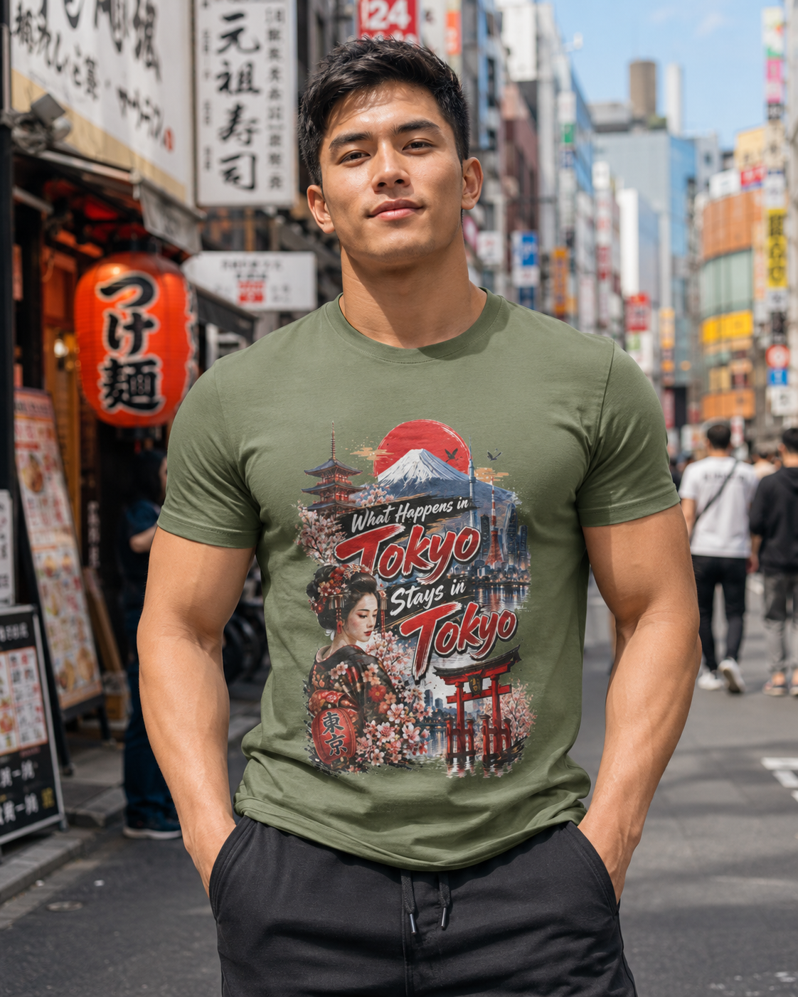 A muscular man wearing an olive green graphic t-shirt with a 'Tokyo Stay in Tokyo' Japanese-themed print, standing on a busy city street.